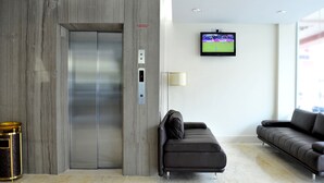 Un vestíbulo moderno con piso de mármol, un sofá, un sillón, un ascensor y un televisor que muestra un evento deportivo.