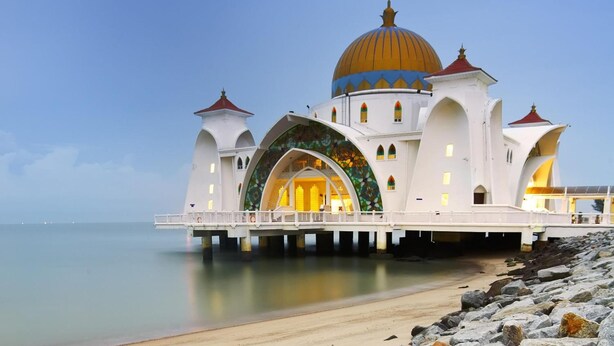 Una mezquita con una gran cúpula dorada y fachada blanca, situada frente a la costa con un muelle que se adentra en el agua.