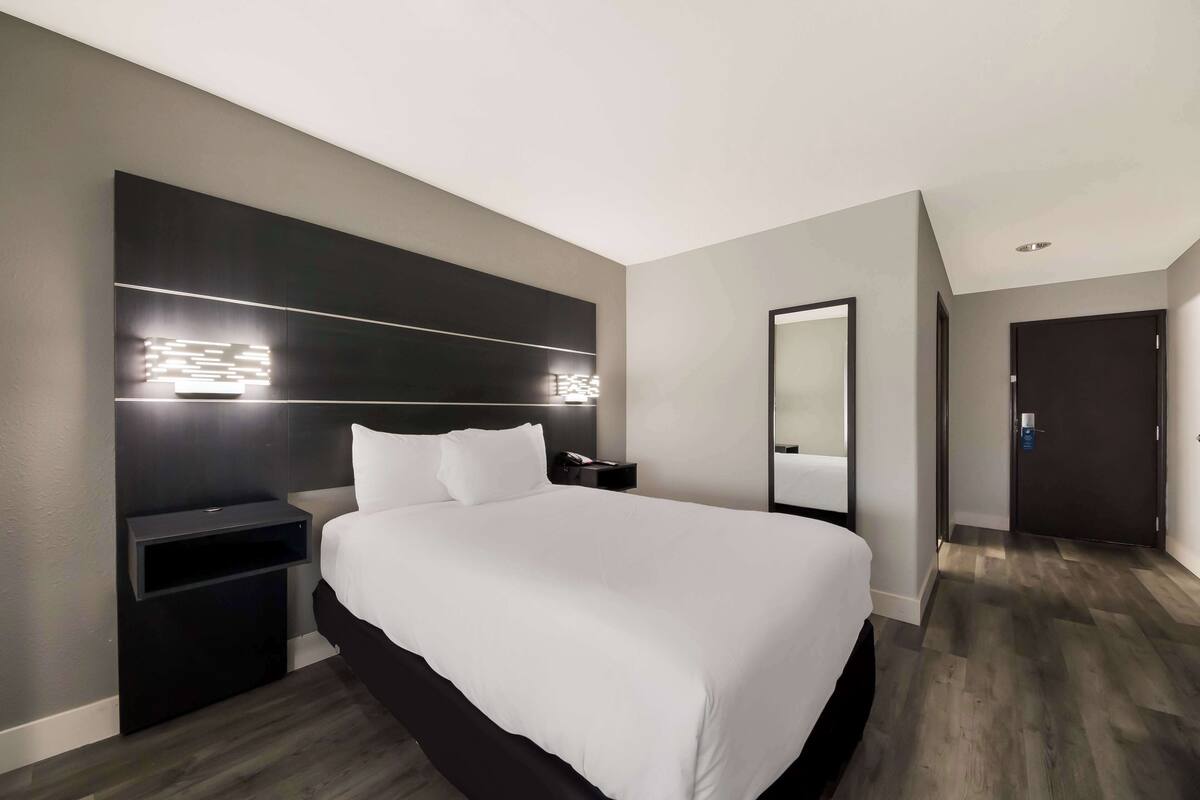A modern hotel room with a large bed, bedside tables, a mirror, and a dark wood floor.