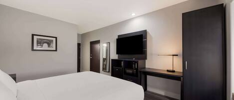 A modern hotel room with a large bed, a flat-screen TV mounted on the wall, a desk with a lamp, and a wardrobe.