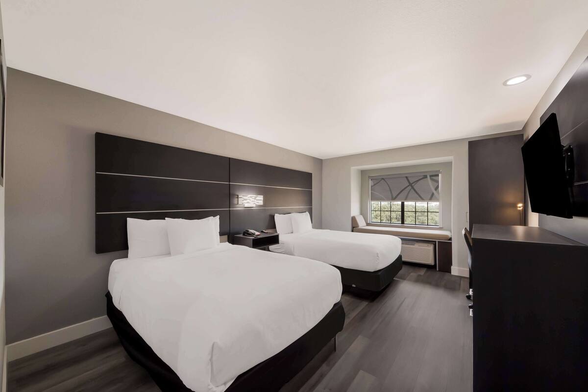 A modern hotel room with two beds, a large dark headboard, a window with a view, and a flat-screen TV.