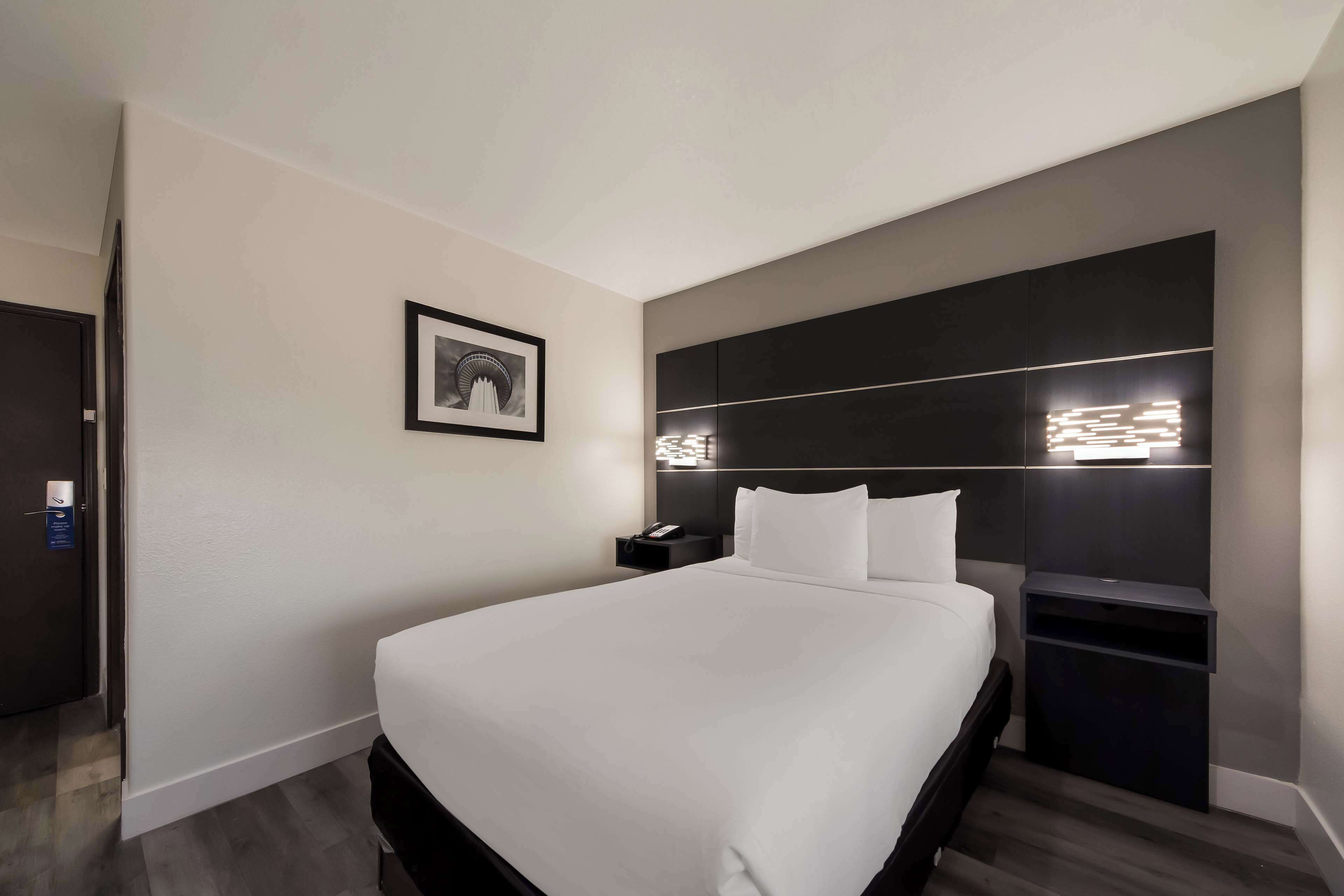 A modern hotel room with a large bed, a bedside table with a lamp, a framed picture on the wall, and a dark headboard with built-in lighting.