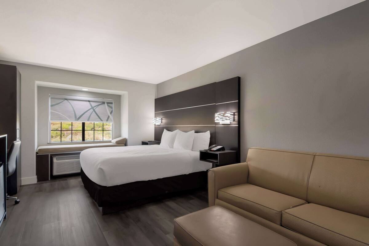 A modern hotel room with a large bed, a sofa, a desk, and a window with a view.
