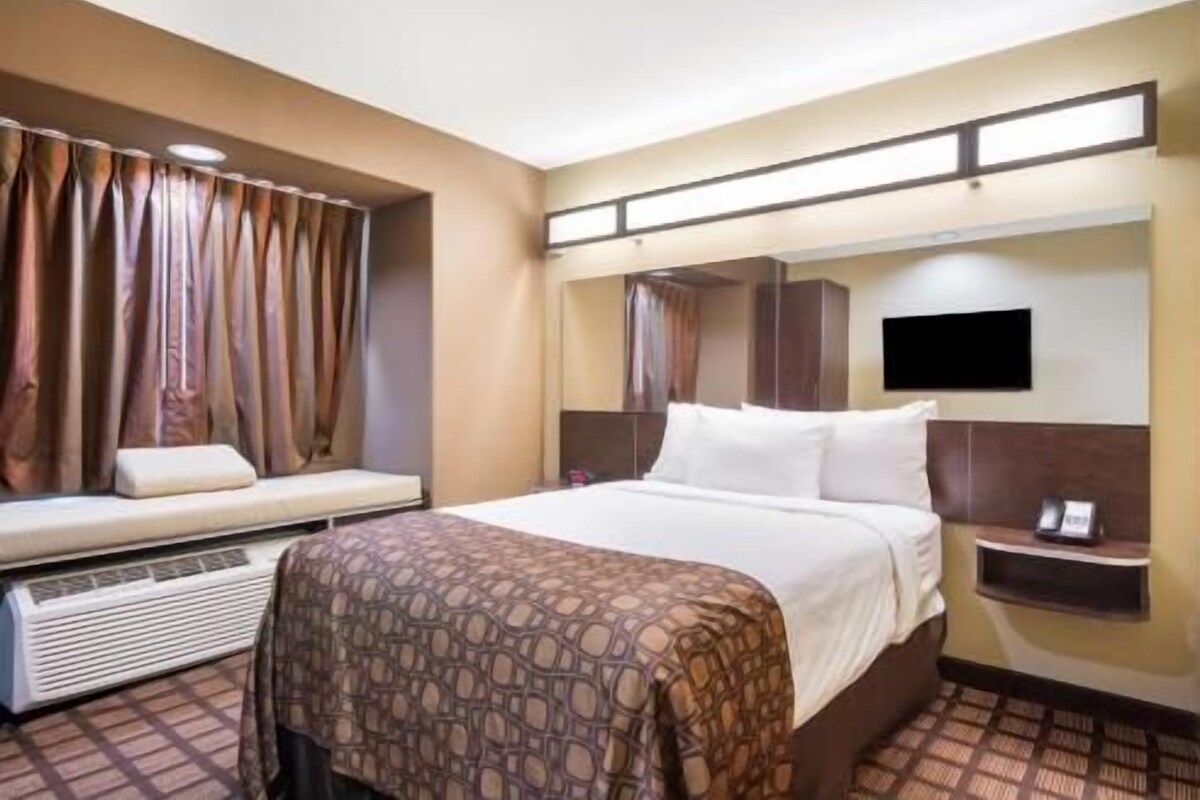 A hotel room with a bed, a bedside table with a phone, a wall-mounted TV, and a window with curtains.