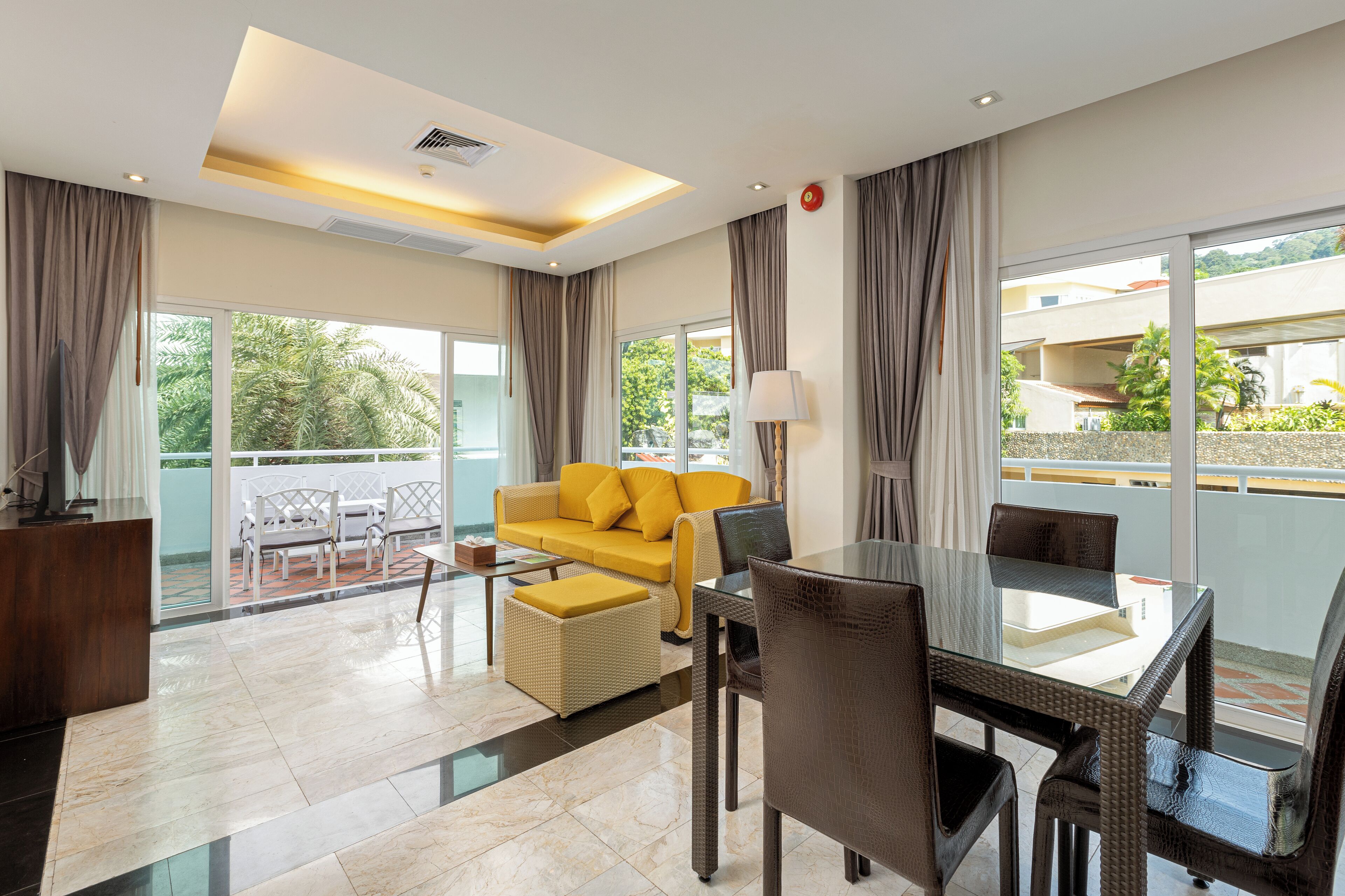 Habitación de hotel moderna con sofá amarillo, mesa de comedor y sillas, ventanas amplias con cortinas y un balcón con mobiliario exterior.