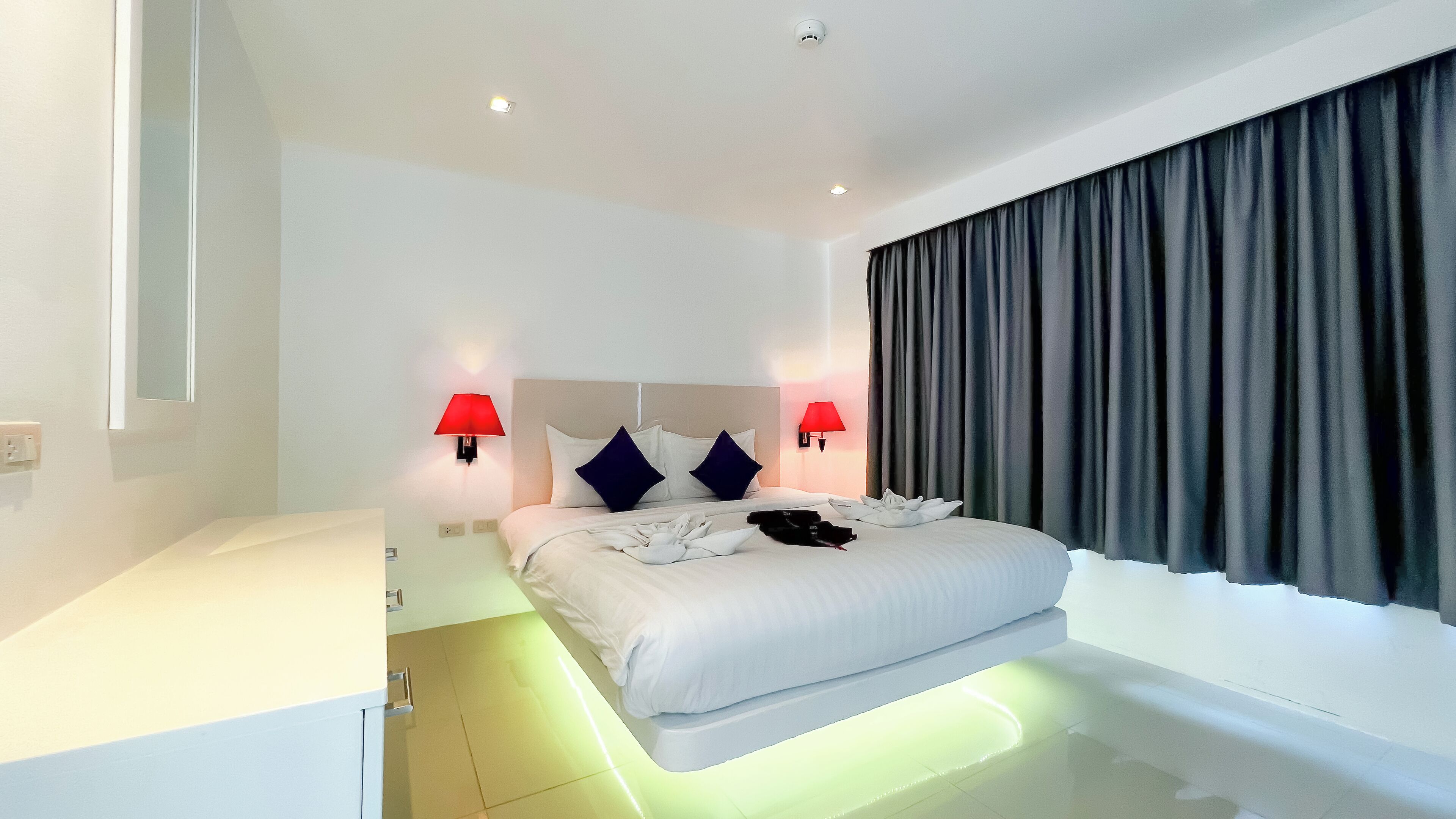 A modern hotel room with a large bed, two red wall lamps, a headboard, and a dark curtain.