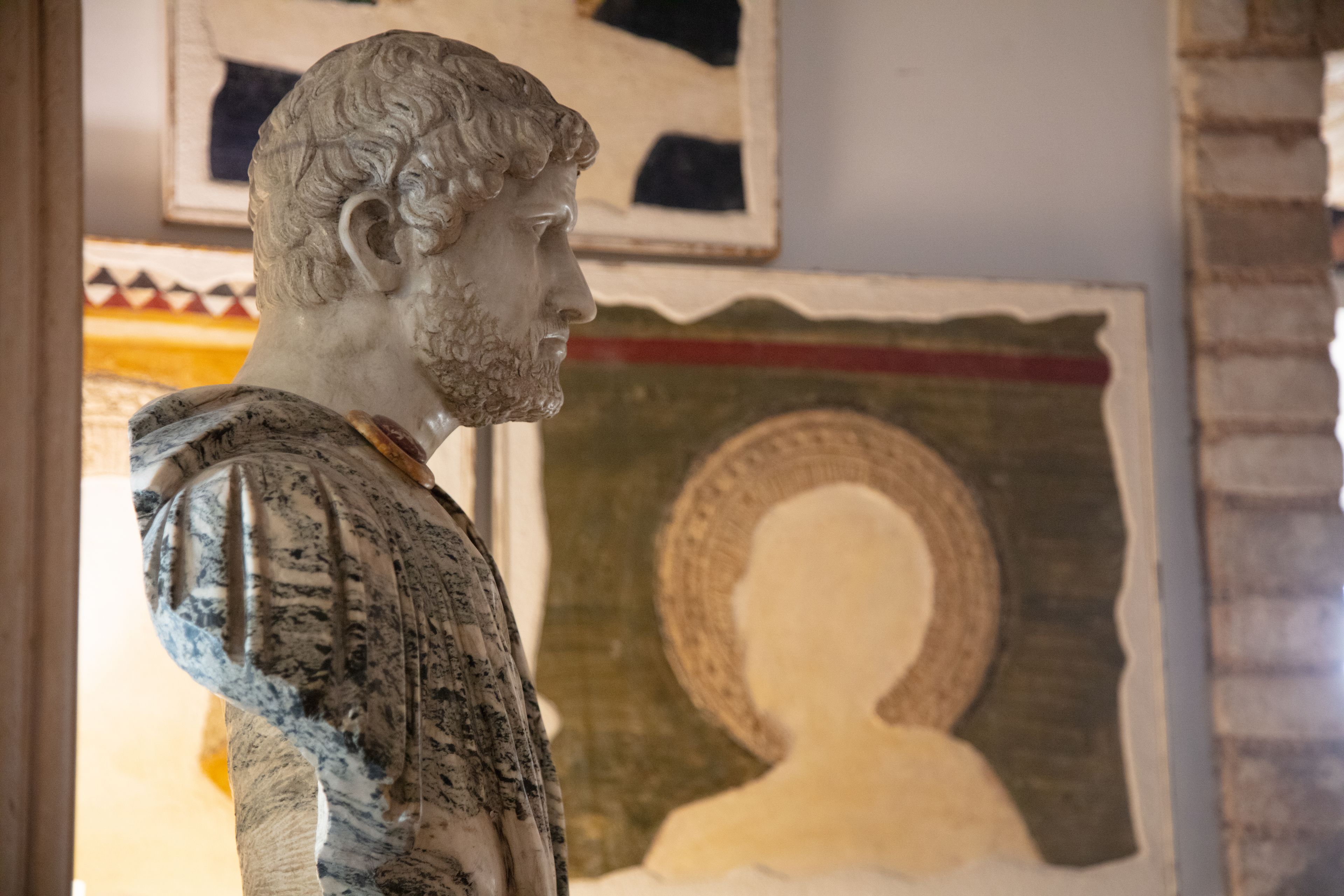 A marble bust sculpture in a room with a framed portrait and a patterned wall.