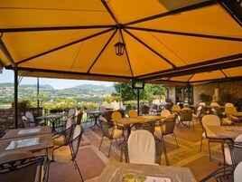 Espaço de refeições ao ar livre com mesas e cadeiras sob um grande guarda-sol, com vista para uma paisagem deslumbrante.