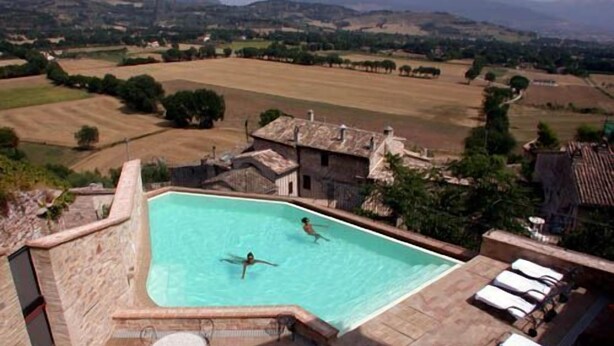 A swimming pool with two people in the water, surrounded by a stone patio with lounge chairs and a table. - Hotel Ristorante La Bastiglia (Spello)