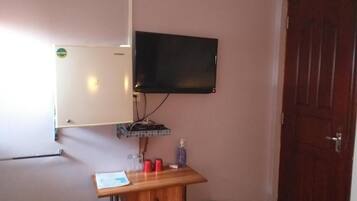 Una habitación con una pizarra, un televisor montado, un escritorio de madera con un cesto de basura y una puerta.