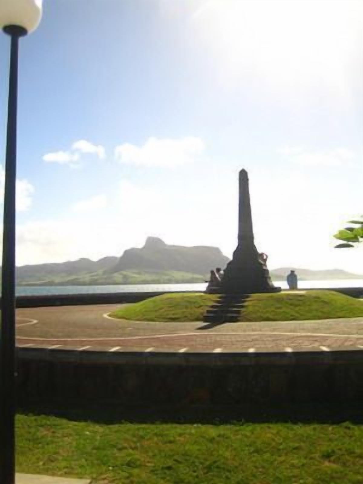 Um monumento com um pilar central, rodeado por uma área gramada circular, e montanhas ao fundo.