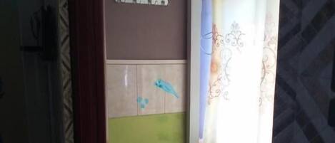 Una habitación pequeña con piso de baldosas, aire acondicionado instalado en la pared, una mesita con una silla y una ventana con cortinas estampadas.