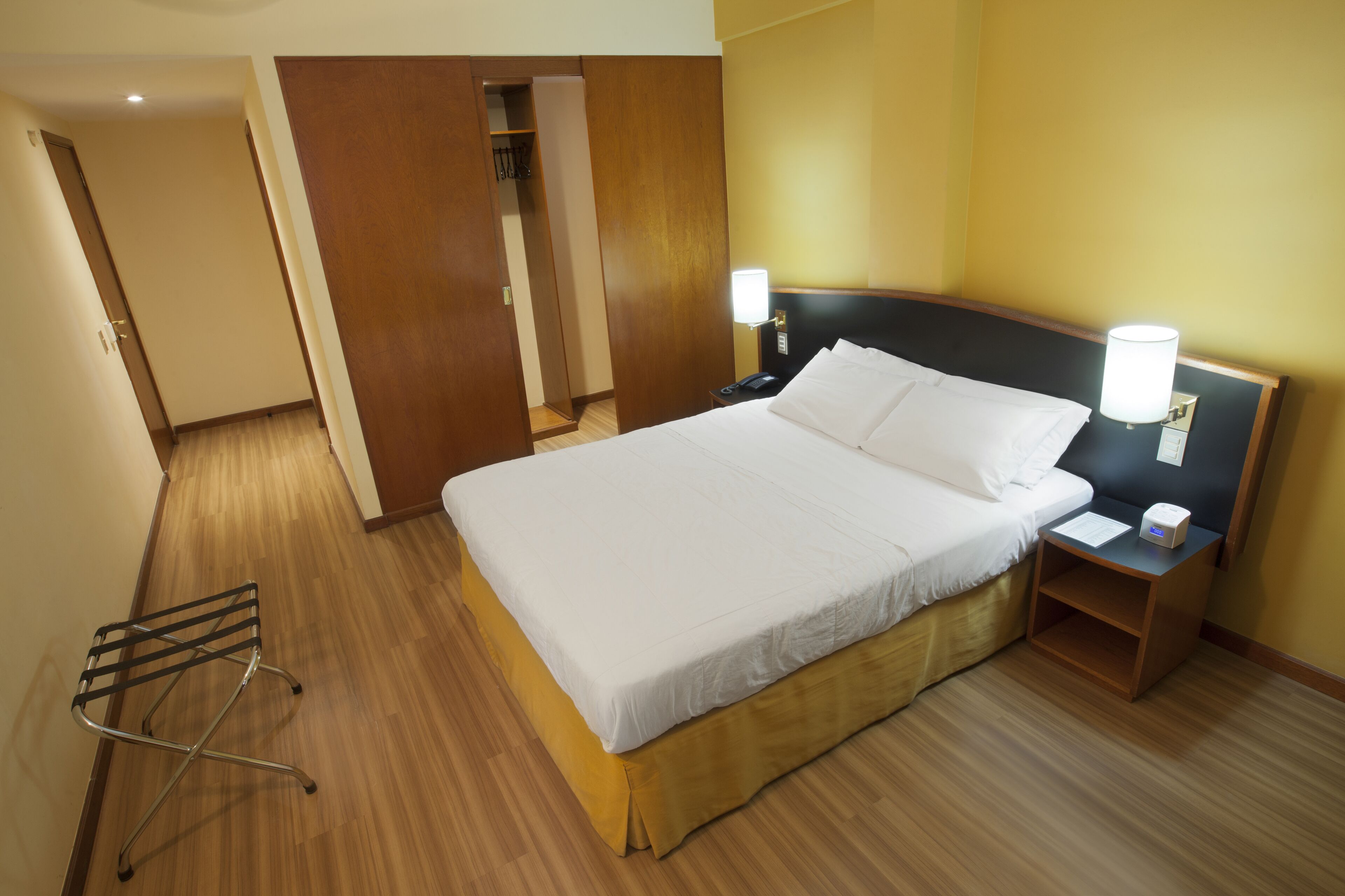 Een hotelkamer met een bed, een nachtkastje met een lamp, een houten vloer en een deur naar een inloopkast.
