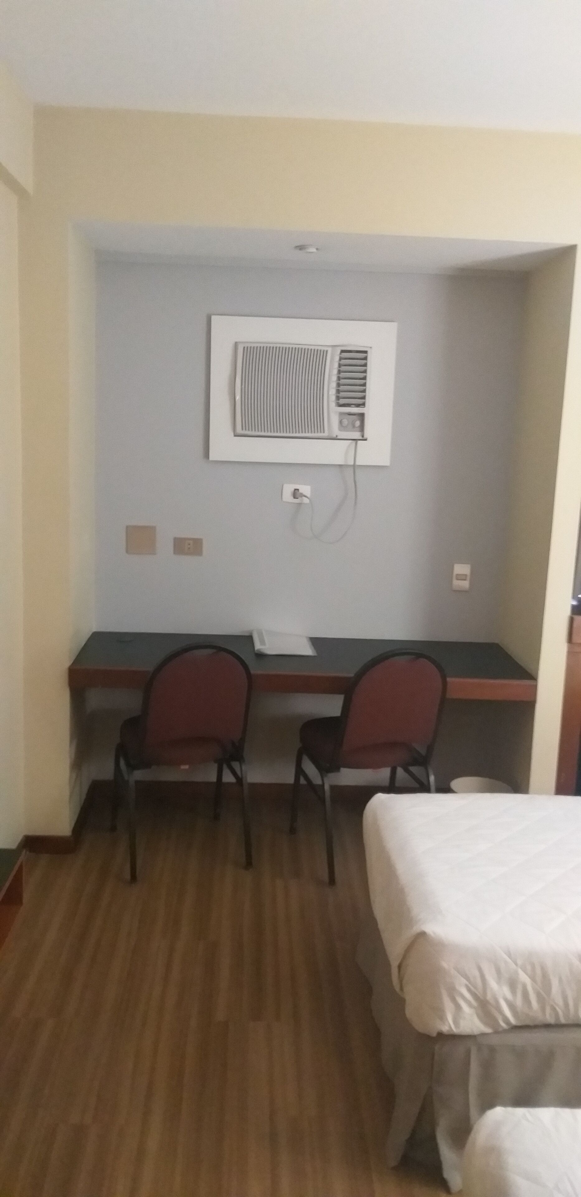 A hotel room with a desk, two chairs, a bed, and an air conditioning unit on the wall.