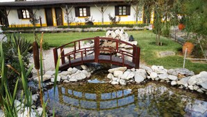 A bridge over a pond with rocks, surrounded by a garden with a house in the background. - A Coutada Hotel Rural (Peniche)