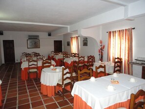 A dining area with multiple round tables, each with white tablecloths and orange sashes, surrounded by wooden chairs.