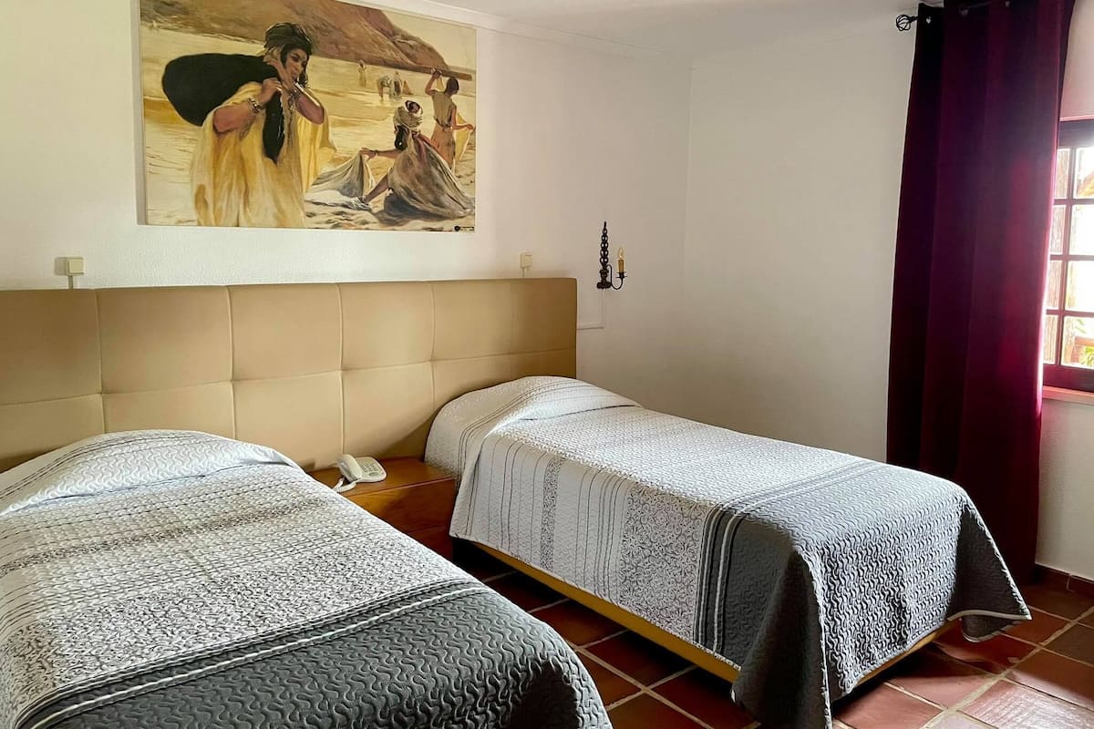 A room with two beds, a painting on the wall, a bedside table with a phone, and a window with curtains.