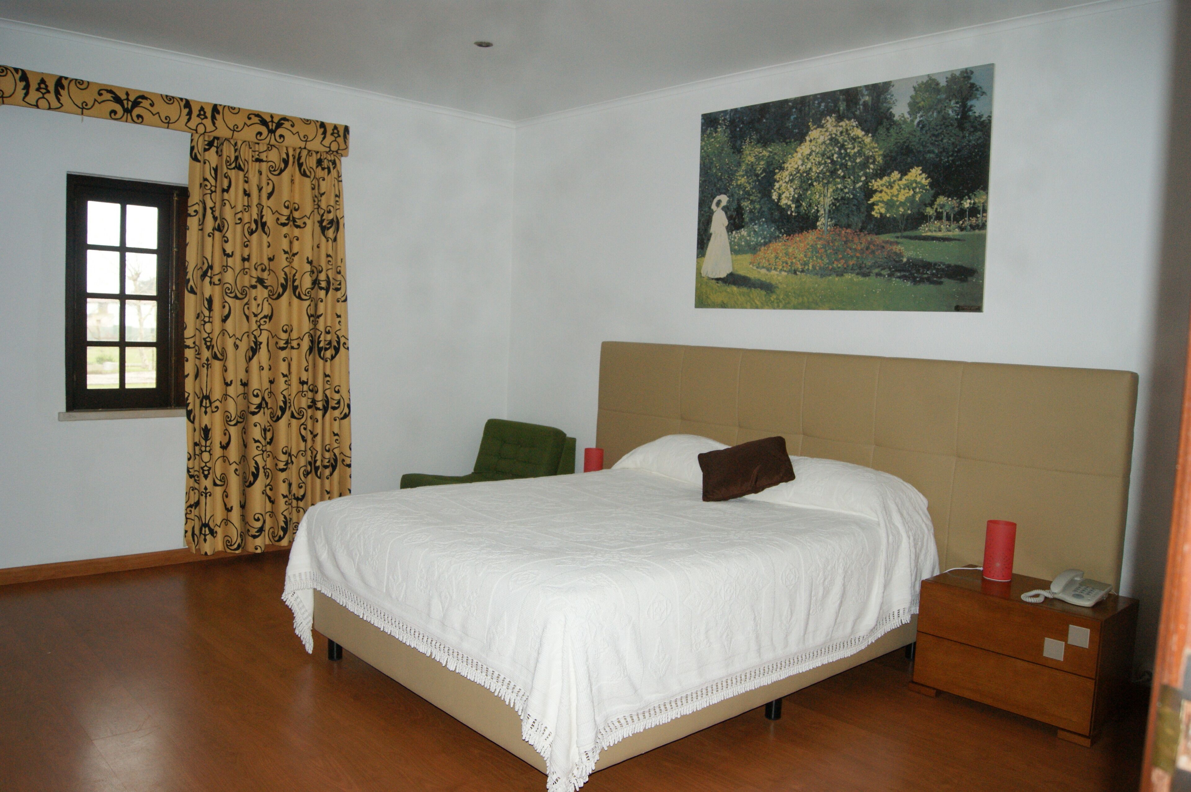 A bedroom with a large bed, a nightstand, a green chair, a window with curtains, and a painting on the wall.
