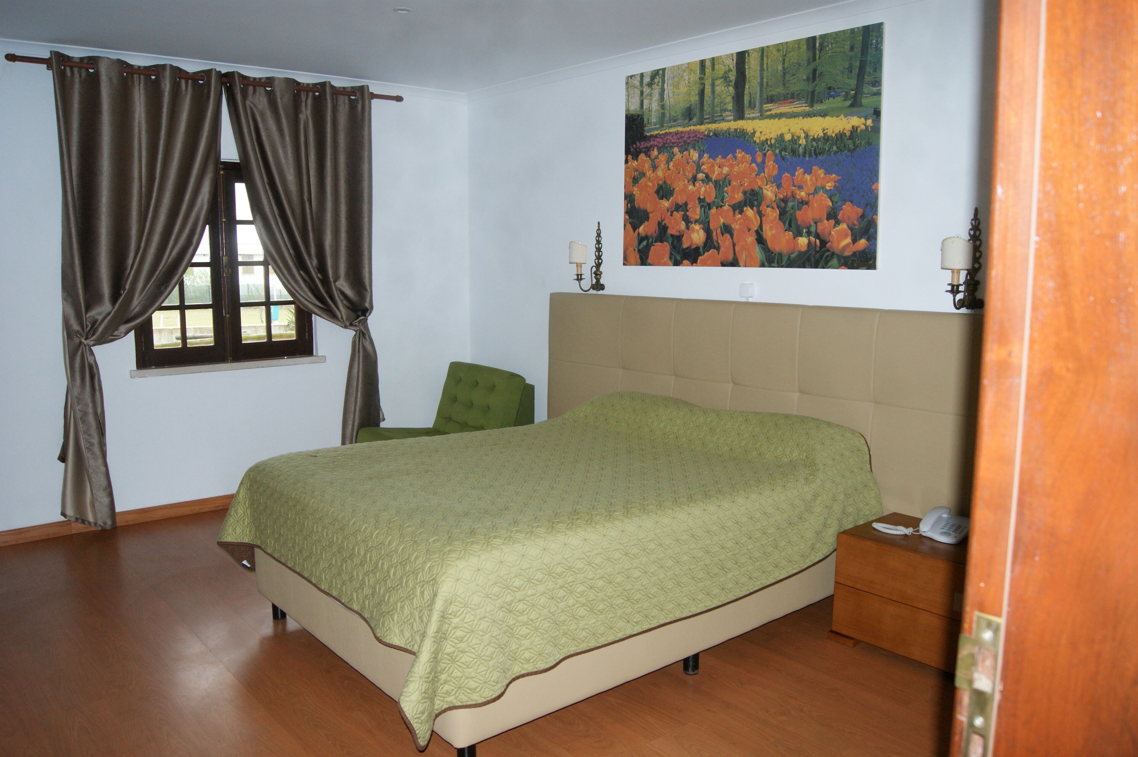 Un dormitorio con cama, una mesita de noche, una silla verde, una ventana con cortinas y un cuadro en la pared.