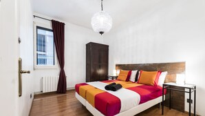 A hotel room with a bed, a wardrobe, a nightstand, a window with curtains, and a chandelier. - Vidre Home - Plaza Real (Barcelona)