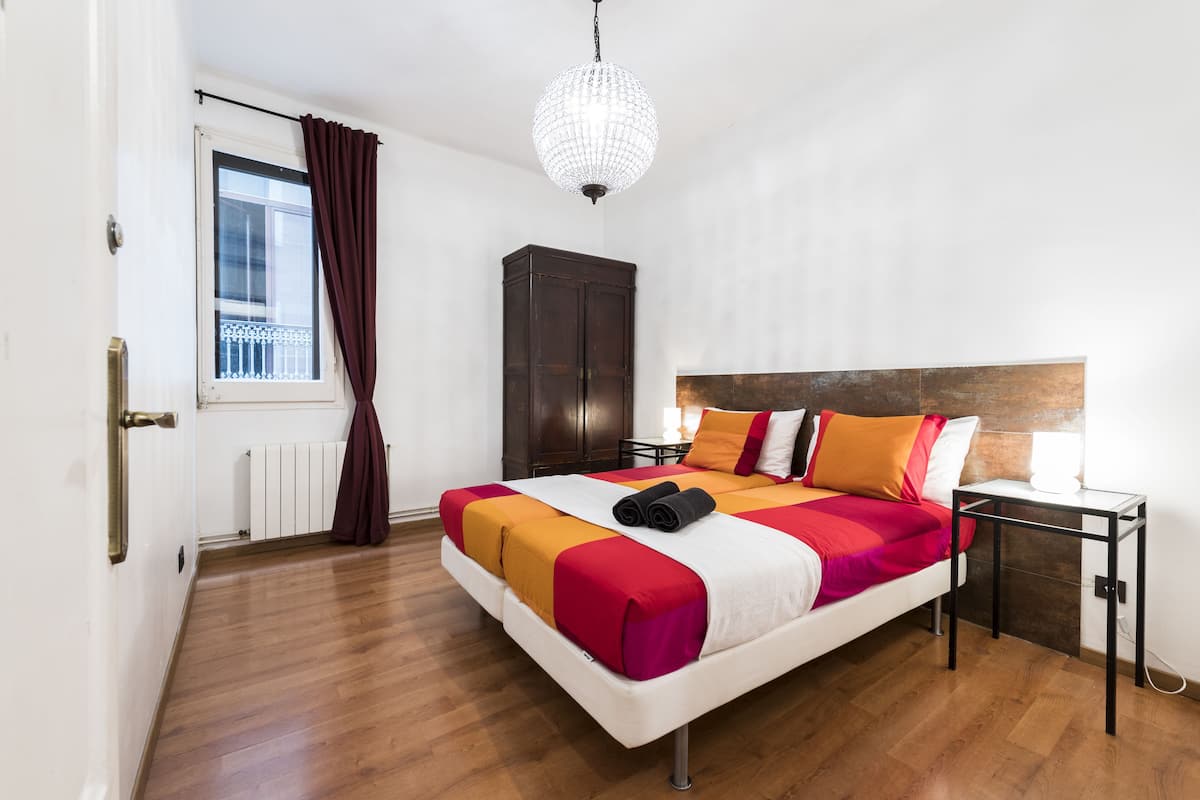 A hotel room with a bed, a wardrobe, a nightstand, a window with curtains, and a chandelier.