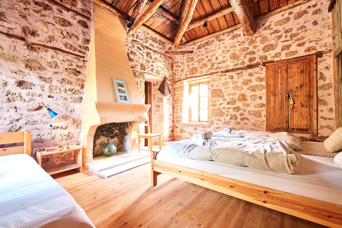 A rustic room with a stone wall, wooden ceiling beams, a bed, a fireplace, and a window.