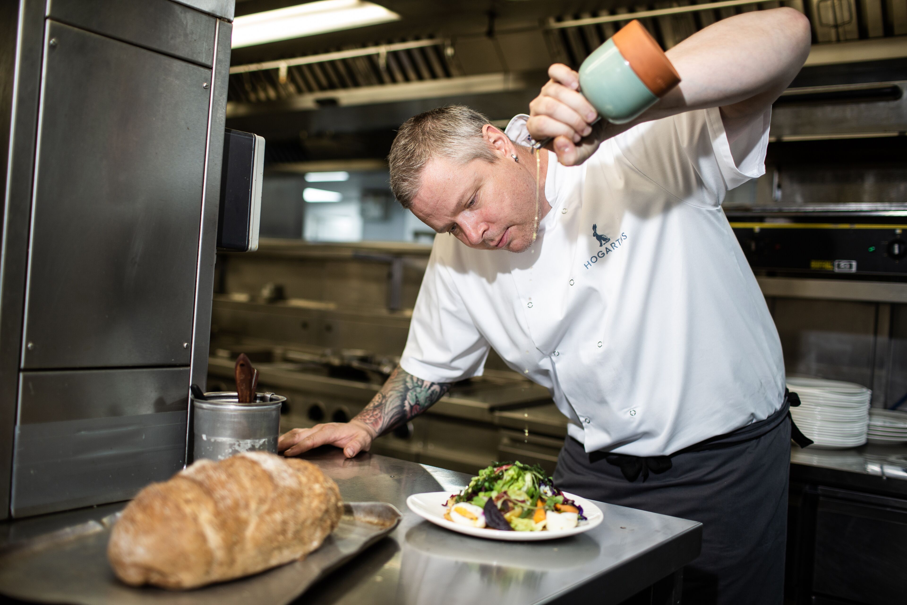 Un chef en una cocina profesional vertiendo salsa de una jarra sobre un plato de comida.