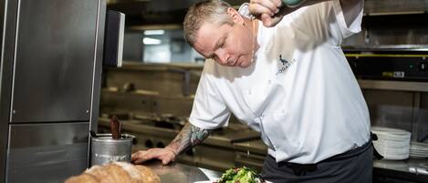 Un chef en una cocina profesional vertiendo salsa de una jarra sobre un plato de comida.