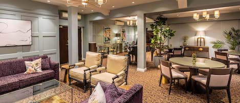 A spacious hotel lobby with a purple sofa, a glass coffee table, and a dining area with a round table and chairs.