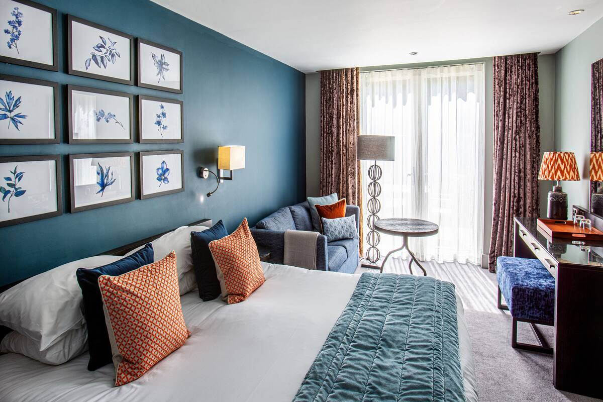 A hotel room with a large bed, a sofa, a small table, and a desk with a chair. There are framed pictures on the wall and a window with curtains.