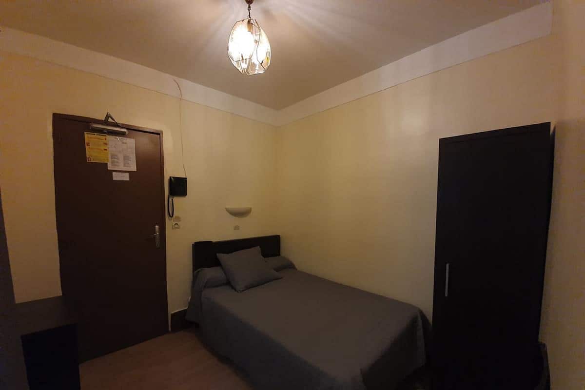 A small, single-bed room with a wooden floor, a dark-colored wardrobe, a bedside lamp, and a ceiling light fixture.