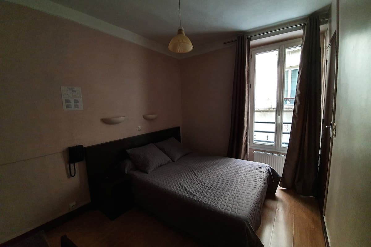 A bedroom with a bed, a hanging light fixture, a window with curtains, and a wall-mounted telephone.