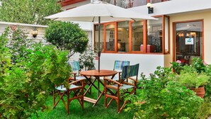 Un patio con mesa y sillas de madera, rodeado de abundante vegetación y un edificio blanco con grandes ventanales.
