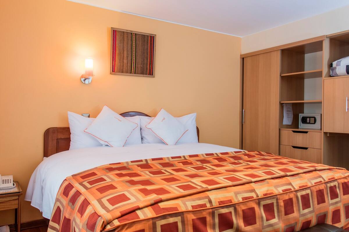 Una habitación de hotel con cama, mesita de noche con teléfono, armario de madera y lámpara fijada a la pared.