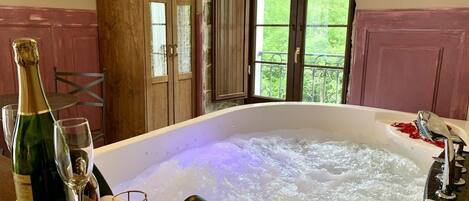 Un jacuzzi con una botella de champán y una copa sobre una mesa de madera, en una habitación con vistas a la vegetación a través de un amplio ventanal.