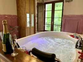 Un jacuzzi con una botella de champán y una copa sobre una mesa de madera, en una habitación con vistas a la vegetación a través de un amplio ventanal.