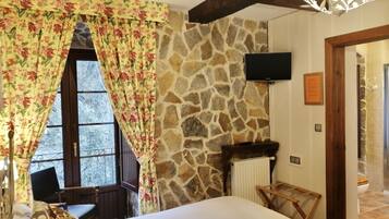 A bedroom with a stone wall, a bed, a wooden chair, a small table, a TV, and a bathroom door.