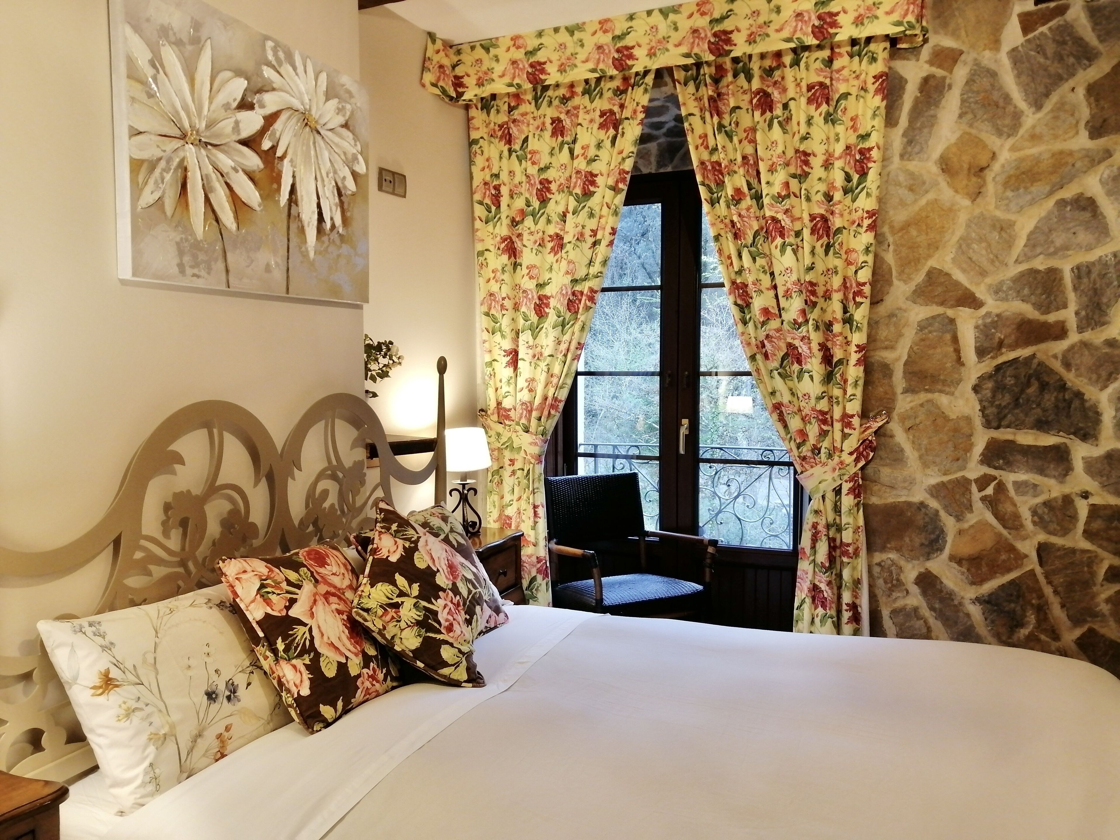 Un dormitorio con cama, cortinas florales, pared de piedra y ventana con vista a árboles.