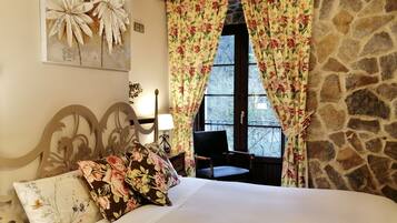 Un dormitorio con cama, cortinas florales, pared de piedra y ventana con vista a árboles.