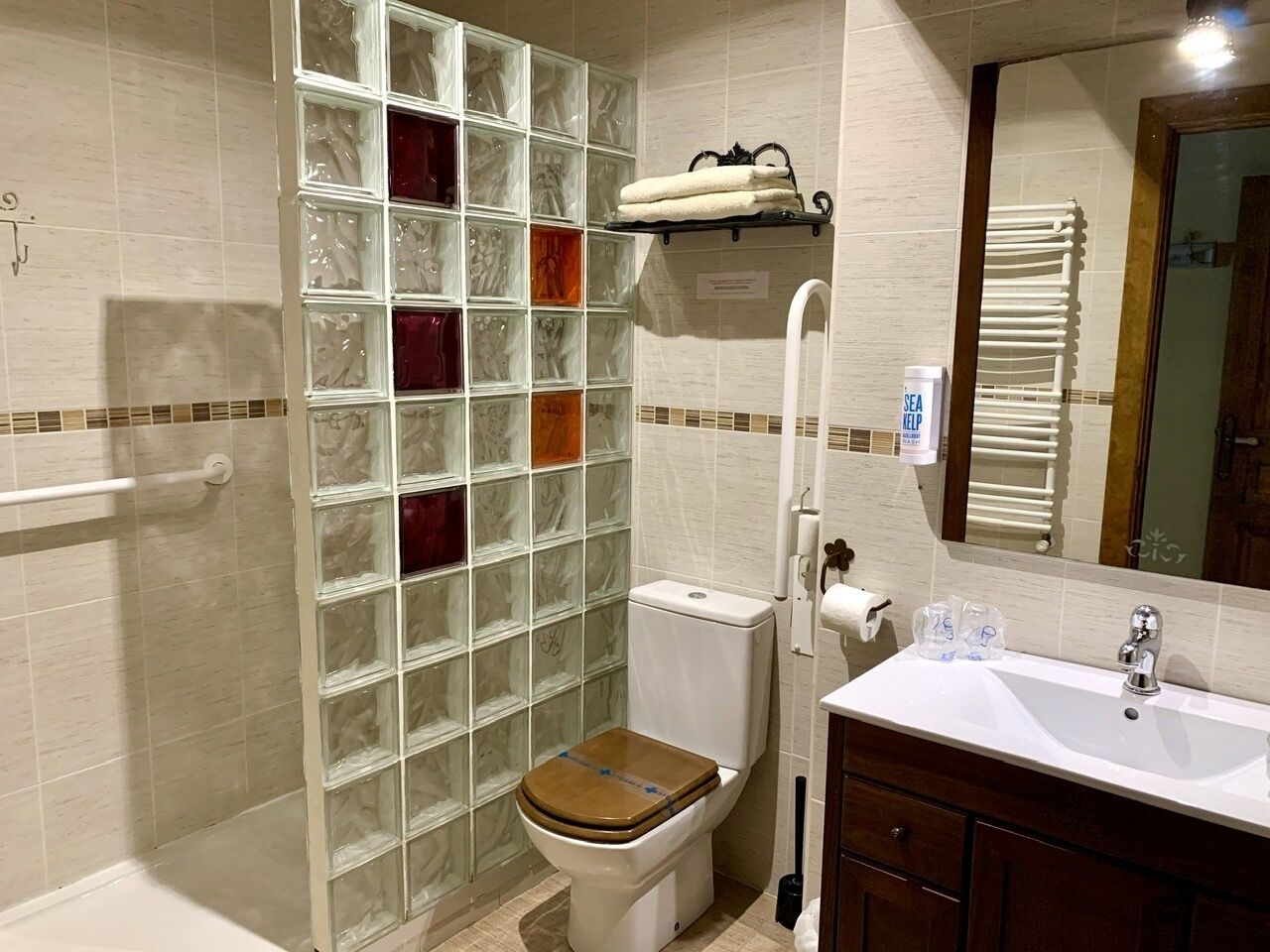 A bathroom with a glass shower enclosure, a toilet with a wooden seat, a white sink, and a mirror above the sink.