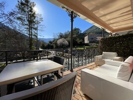 Un patio al aire libre con una mesa y sofá blanco, rodeado por una barandilla metálica, con vistas a un paisaje pintoresco con árboles y una montaña.