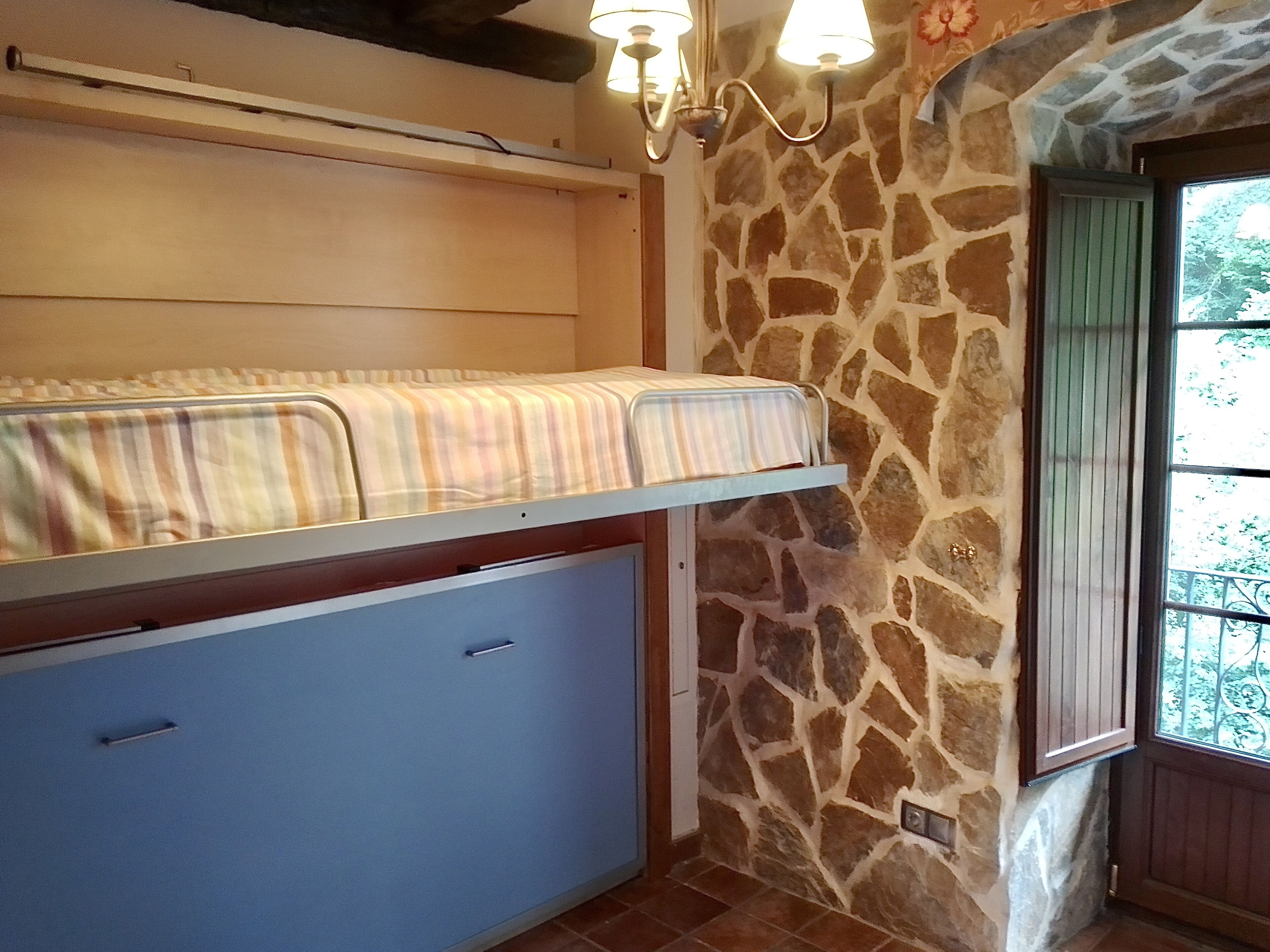 A bunk bed with a striped blanket, a stone wall, a window with a view, and a light fixture.