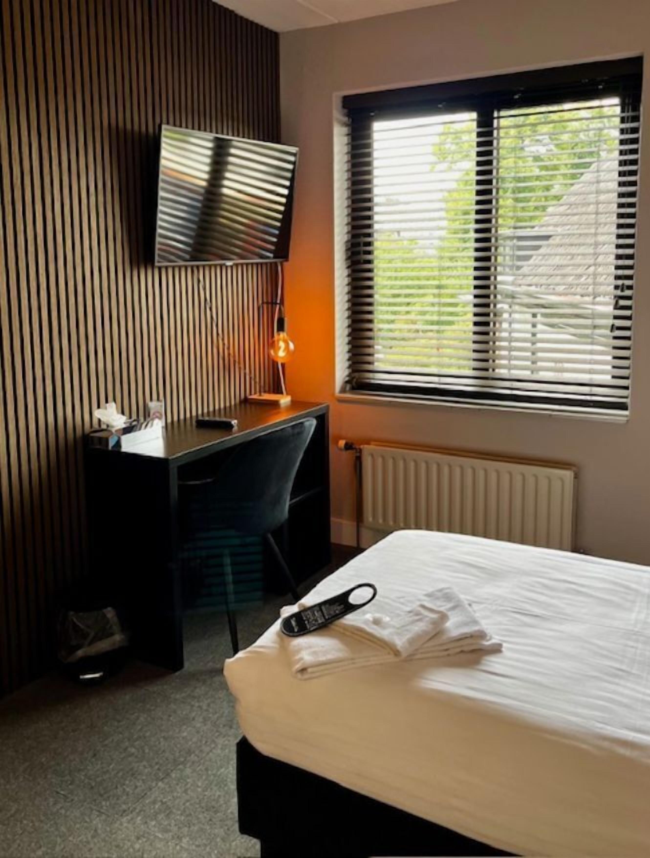A hotel room with a bed, a desk with a chair, a TV mounted on the wall, and a window with blinds.