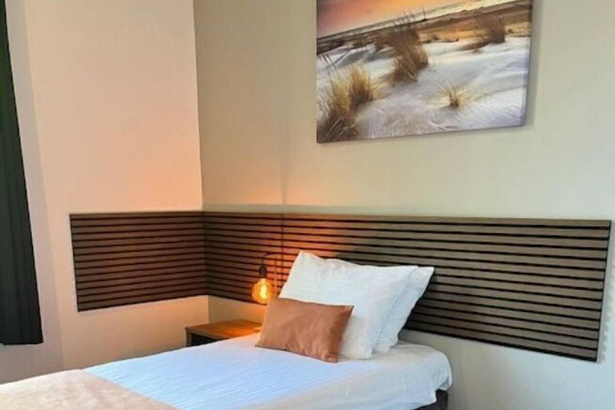 A hotel room with a bed, a nightstand, a painting of a sunset, and a dark carpet.
