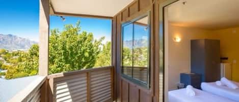 Un balcón con vistas a montañas y vegetación, un dormitorio con cama y mesita de noche, y una barandilla de madera.