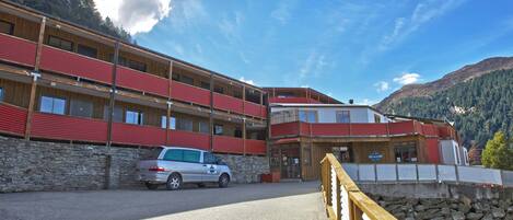 A multi-story building with a stone facade and red accents, featuring a parking area and a wooden deck overlooking a mountainous landscape.
