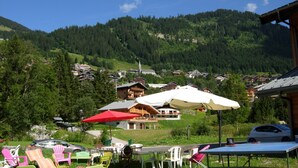A mountain village with traditional wooden houses, outdoor seating with umbrellas, and a view of the surrounding forested hills.