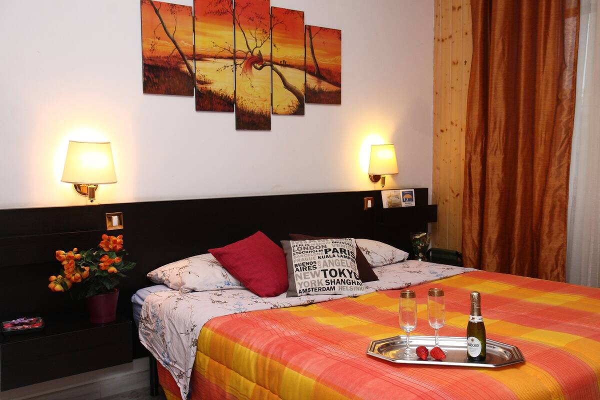 A hotel room with a bed, two wall-mounted lamps, a nightstand with a plant, a tray with champagne glasses and strawberries, and a city-themed pillow.