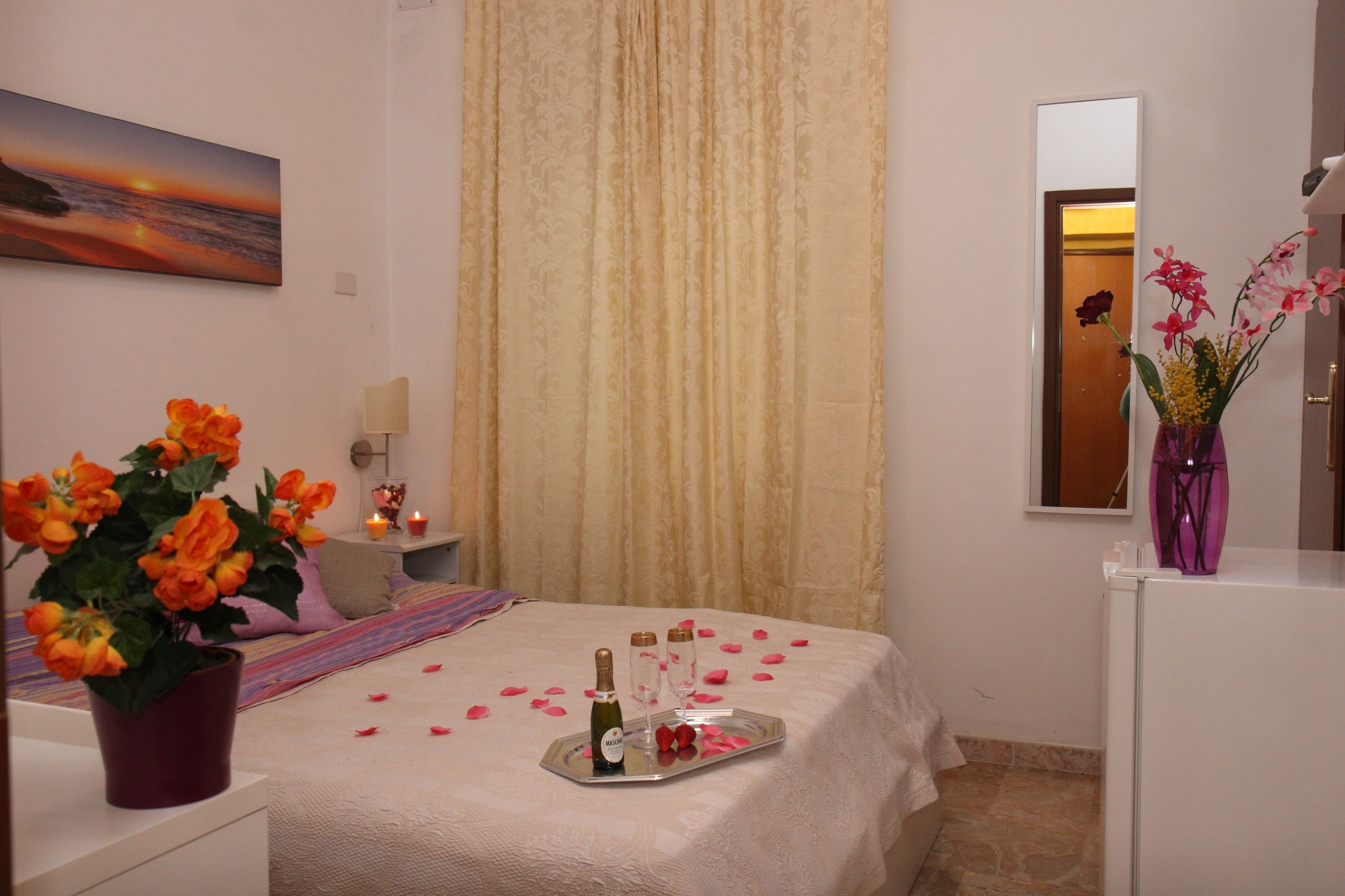 A hotel room with a bed, a potted plant, a tray with a bottle and glasses, and a vase with flowers.