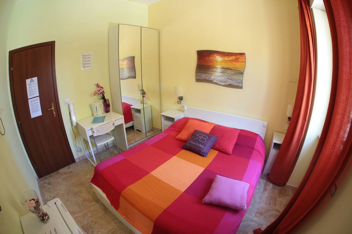 A bedroom with a bed, a desk, a mirror, and a painting on the wall.